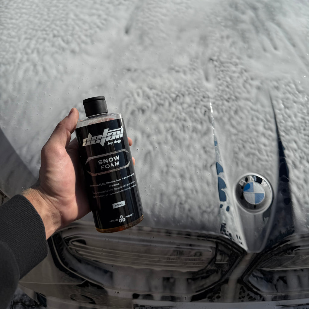 Hand holding a bottle of snow foam in front of a BMW car with snow on the ground.